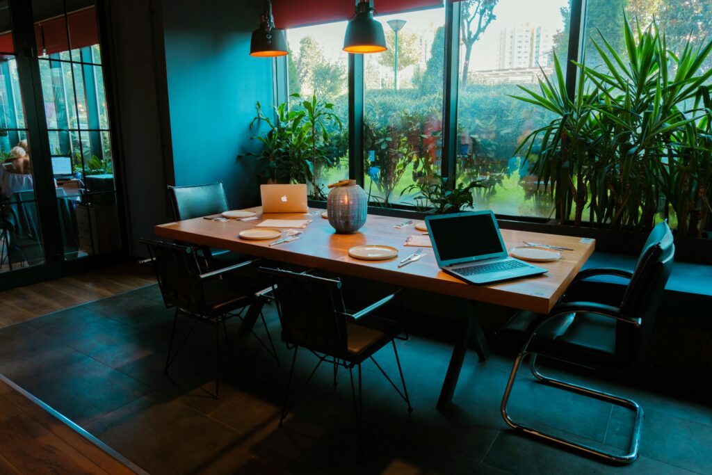 Hakkımızda 2 A modern dining room setup with laptops, plates, and lush plants by large windows.